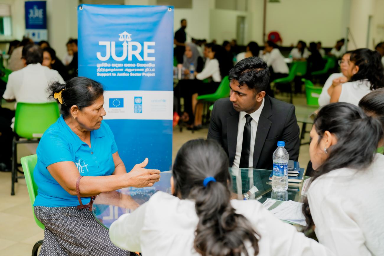 A group discussion around a table, with a woman in blue speaking passionately to others.